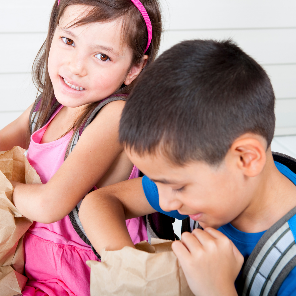 Two children; one girl in a pink dress smiling at the camera and one boy in a blue shirt reaching into a brown lunch bag.
